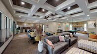 Located at 101 Hollow Tree Ln in Houston, TX, this single-floor community clubhouse is designed to serve as a dynamic social and functional hub. The centerpiece of the space is a generously sized open-concept great room featuring a striking coffered ceiling with dark wood inlays and recessed lighting that adds architectural depth and warmth. This area seamlessly connects a comfortable lounge with leather sofas, a game room with a full-size pool table, and a stone fireplace that anchors the space with a classic touch. Multiple dining tables with colorful modern chairs provide ample seating throughout, while large windows and French doors invite abundant natural light and outdoor views. The full community kitchen is well-appointed with white cabinetry, granite countertops, a wood-plank backsplash, stainless steel appliances, a central island with bar seating, and a built-in wine rack. A dedicated lounge nook features vibrant teal accent walls, a wall-mounted TV, and a sliding barn door, creating a cozy media retreat. Additional areas include a bright waiting or coffee nook with orange leather chairs and garden views, a small private office or media room with a wall-mounted TV and workstation, and a welcoming entryway with colorful accent seating and local Houston-themed artwork throughout.