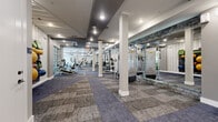 Located at 3964 Watercourse Dr in Fort Worth, TX, this well-appointed fitness center offers a substantial single-floor layout designed to accommodate a full range of workout needs. The space features a clean, modern aesthetic with neutral-toned walls, recessed lighting, and stylish patterned carpet tiles throughout that add visual texture while providing a comfortable underfoot surface. White structural columns punctuate the open floor plan, contributing to the architectural character of the space without impeding movement between equipment zones. Large floor-to-ceiling mirrors with stone-accented trim line one wall, enhancing the sense of openness and allowing users to monitor their form during workouts. The cardio area is equipped with treadmills, ellipticals, and stair climbers, positioned near large windows and glass garage-style doors that invite abundant natural light and views of the surrounding greenery. The strength training zone features a variety of cable machines, weight stations, and free weights including a full dumbbell rack with an adjustable bench. A dedicated accessories wall organizes stability balls, resistance bands, and other functional fitness tools. Ceiling fans and recessed lighting ensure comfort throughout. The overall ambiance is bright, airy, and purpose-driven, making this fitness center a comprehensive and inviting facility.