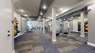 Located at 3964 Watercourse Dr in Fort Worth, TX, this well-appointed fitness center offers a substantial single-floor layout designed to accommodate a full range of workout needs. The space features a clean, modern aesthetic with neutral-toned walls, recessed lighting, and stylish patterned carpet tiles throughout that add visual texture while providing a comfortable underfoot surface. White structural columns punctuate the open floor plan, contributing to the architectural character of the space without impeding movement between equipment zones. Large floor-to-ceiling mirrors with stone-accented trim line one wall, enhancing the sense of openness and allowing users to monitor their form during workouts. The cardio area is equipped with treadmills, ellipticals, and stair climbers, positioned near large windows and glass garage-style doors that invite abundant natural light and views of the surrounding greenery. The strength training zone features a variety of cable machines, weight stations, and free weights including a full dumbbell rack with an adjustable bench. A dedicated accessories wall organizes stability balls, resistance bands, and other functional fitness tools. Ceiling fans and recessed lighting ensure comfort throughout. The overall ambiance is bright, airy, and purpose-driven, making this fitness center a comprehensive and inviting facility.