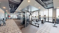 Located at 615 E Morehead St in Charlotte, NC, this modern fitness center offers a generous, open floor plan designed to accommodate a comprehensive range of workout needs. The space is defined by its floor-to-ceiling windows along multiple walls, flooding the interior with natural light and framing striking views of the Charlotte skyline. The industrial-inspired design features exposed ductwork and structural elements overhead, contrasted by sleek tray ceilings with recessed lighting that create distinct zones throughout the facility. A dedicated strength training area is equipped with power racks, weight benches, cable machines, and an extensive free weight selection. A separate cardio zone lines the window-facing perimeter, housing treadmills, ellipticals, stationary bikes, and stair climbers. Glass-walled studio spaces with wood-look flooring provide room for stretching, functional training, and group exercise. A vibrant graphic mural accent wall adds an energetic aesthetic element, while water fountains and wall-mounted TVs enhance convenience. The flooring transitions thoughtfully between rubber, carpet tile, and hardwood-style surfaces to suit each functional zone, contributing to a well-organized and motivating training environment.