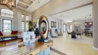This elegantly appointed single-floor clubhouse serves as a premier amenity space designed for comfort, productivity, and community. The grand entrance lobby immediately impresses with its soaring coffered ceilings, large-format light tile flooring, and curated décor, including a striking bronze sculptural centerpiece displayed on a reception console. Abundant natural light flows in through tall, multi-pane windows framed by floor-length drapery, enhancing the bright, welcoming atmosphere throughout.

The expansive main lounge anchors the space with a dramatic floor-to-ceiling stone fireplace, a wall-mounted television with built-in display niches, and generous seating arrangements accented by colorful area rugs and lush tropical plants. A pendant chandelier adds warmth above, while large glass doors invite views to the outdoors.

Adjacent to the lounge, a dedicated dining and gathering area features contemporary round tables and modern seating, alongside a built-in coffee station set within an arched niche. A separate business center provides additional workspace, clearly marked and conveniently accessible.

The well-appointed kitchen offers dark wood cabinetry, granite countertops, a mosaic tile backsplash, stainless steel appliances, and a bar-height peninsula with seating. A glass-walled fitness center, visible from the corridor, is equipped with cardio machines and ceiling fans, completing this thoughtfully designed community facility.