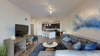This single-floor unit showcases a thoughtfully designed open-concept layout that seamlessly connects the living area and kitchen into one cohesive living space. The living room is anchored by a plush grey sectional sofa paired with a round white coffee table and a patterned navy area rug, creating an inviting and modern atmosphere. A large flat-screen television is mounted on a sleek media console along one wall, while contemporary wall art adds a vibrant touch of color to the otherwise neutral palette.

The kitchen features dark espresso cabinetry contrasted by a bright white island with bar seating, offering an ideal spot for casual dining or entertaining. Pendant lighting hangs above the island, complementing the track lighting overhead and recessed fixtures that illuminate the entire space. Stainless steel appliances add a polished, modern finish to the kitchen.

Hardwood-style flooring runs throughout the main living areas, contributing warmth and continuity to the design. The high, white ceilings amplify the sense of openness and airiness. Overall, the space presents a clean, contemporary aesthetic with a well-coordinated blue and grey color scheme.