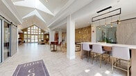 The 803 Jordan clubhouse in Naperville, Illinois is a thoughtfully designed community space that combines contemporary elegance with welcoming functionality. The expansive single-floor layout is anchored by a dramatic cathedral-ceiling entryway, where floor-to-ceiling windows flood the interior with natural light and offer views of the surrounding greenery. Light-toned large-format tile flooring extends throughout the main areas, contributing to the bright and airy atmosphere.

The open main hall serves as a central hub, featuring a bar-height counter with seating, pendant lighting, and a striking herringbone wood accent wall bearing the building's branding. Adjacent areas transition seamlessly into distinct activity zones: a comfortable lounge with plush seating, a wall-mounted flat-screen TV, and curated artwork; a dining area with a glass-top table and bold blue upholstered chairs; and a game room complete with a billiards table.

A standout feature is the floor-to-ceiling stone fireplace with a reclaimed wood mantle, adding warmth and a rustic focal point to the social area. Sliding barn-style doors lead to a private meeting or flex room, while geometric-patterned carpeting defines the lounge zones. A geometric gold chandelier and industrial-style pendant lights add refined finishing touches throughout this well-appointed community clubhouse.