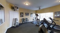 This fitness room offers a practical and well-organized space for exercise and physical wellness. The room features warm yellow walls, white trim, and dark carpet flooring that creates a focused workout atmosphere. Natural light streams in through large windows with vertical blinds, brightening the space and adding an open feel. A ceiling fan provides additional ventilation and comfort during workouts.

The space is equipped with a solid selection of fitness equipment, including a treadmill, an elliptical machine, and a multi-function cable weight station, catering to both cardio and strength training needs. A dedicated free weights rack holds a range of dumbbells, making it easy to incorporate versatile strength exercises into any routine. A wall-mounted television is positioned for easy viewing, allowing users to stay entertained while working out.

Motivational and fitness-themed artwork lines the walls, contributing to an energizing environment. Two chairs are available as a rest area, offering a place to recover between sets. The layout of the room is thoughtfully arranged to allow comfortable movement between equipment. Overall, this fitness room provides a functional and welcoming environment well-suited for daily exercise routines.