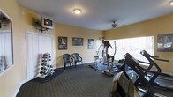 This fitness room offers a practical and well-organized space for exercise and physical wellness. The room features warm yellow walls, white trim, and dark carpet flooring that creates a focused workout atmosphere. Natural light streams in through large windows with vertical blinds, brightening the space and adding an open feel. A ceiling fan provides additional ventilation and comfort during workouts.

The space is equipped with a solid selection of fitness equipment, including a treadmill, an elliptical machine, and a multi-function cable weight station, catering to both cardio and strength training needs. A dedicated free weights rack holds a range of dumbbells, making it easy to incorporate versatile strength exercises into any routine. A wall-mounted television is positioned for easy viewing, allowing users to stay entertained while working out.

Motivational and fitness-themed artwork lines the walls, contributing to an energizing environment. Two chairs are available as a rest area, offering a place to recover between sets. The layout of the room is thoughtfully arranged to allow comfortable movement between equipment. Overall, this fitness room provides a functional and welcoming environment well-suited for daily exercise routines.