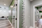 This contemporary single-floor office space is thoughtfully designed to support professional collaboration and productivity. The layout centers around a well-appointed conference room, clearly identified by bold three-dimensional lettering mounted vertically on a sage green accent wall — a striking design detail that sets the tone for the entire space. Entry into the conference area is through glass-paneled doors framed in crisp white trim, lending an open yet defined sense of separation between zones.

The conference room itself features a large rectangular dark-topped table surrounded by a mix of modern black and white chairs, accommodating a sizeable group for meetings or presentations. A wall-mounted flat-screen display serves as a focal point on one wall, while geometric patterned wallpaper adds texture and visual interest. Pendant globe lighting overhead provides warm, focused illumination, and a black-and-white framed art piece completes the polished aesthetic.

Adjacent to the conference room is a casual breakout or collaboration nook, featuring a wall-mounted bar-height counter paired with sleek dark bar stools. Two industrial-style diamond pendant lights hang above, and a bold geometric wallpaper creates a dynamic backdrop. Throughout the space, wood-look plank flooring ties the rooms together with warmth and continuity, while the interplay of soft gray and green tones creates a cohesive, professional ambiance.