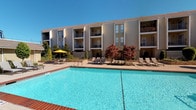 This amenity space offers residents two distinct areas designed for recreation and fitness. The outdoor pool area is a standout feature, showcasing a generously sized, crystal-clear swimming pool with brick-trimmed edges. The surrounding deck is furnished with lounge chairs and a shaded seating area beneath a yellow umbrella, creating an inviting retreat. The multi-story building backdrop, adorned with private balconies and mature ornamental trees, adds a pleasant sense of enclosure to the space.

The fitness center is bright and thoughtfully designed, featuring light-toned herringbone-patterned flooring, recessed lighting, and a ceiling fan for comfort. Floor-to-ceiling mirrors line the walls, amplifying the sense of space and allowing for form checks during workouts. Large windows and sliding glass doors provide natural light and direct views of the pool area. The gym is well-equipped with multiple treadmills, ellipticals, cable machines, adjustable benches, a full dumbbell rack, and medicine balls. Floating wooden shelves offer organized storage for rolled towels and accessories, adding a polished, functional touch to the overall atmosphere.