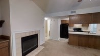 This single-floor space is defined by its open-concept layout, where the living area and kitchen flow seamlessly together. The living area is anchored by a wood-burning fireplace framed in white-trimmed tile, adding a classic focal point to the room. Vaulted ceilings rise above the space, creating an airy, expansive feel enhanced by neutral-toned carpet that extends throughout. The kitchen is positioned adjacent to the living area and features dark wood cabinetry, a black refrigerator, and a black range, all set against light countertops that provide contrast. The layout allows for easy interaction between the kitchen and living space. A hallway visible from the main area leads toward additional rooms, with light-colored flooring transitioning at the entrance. Overhead ventilation is integrated into the ceiling, and recessed lighting provides practical illumination throughout. The overall ambiance is functional and straightforward, with the fireplace and vaulted ceiling serving as the primary architectural highlights of the space.