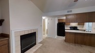This single-floor space is defined by its open-concept layout, where the living area and kitchen flow seamlessly together. The living area is anchored by a wood-burning fireplace framed in white-trimmed tile, adding a classic focal point to the room. Vaulted ceilings rise above the space, creating an airy, expansive feel enhanced by neutral-toned carpet that extends throughout. The kitchen is positioned adjacent to the living area and features dark wood cabinetry, a black refrigerator, and a black range, all set against light countertops that provide contrast. The layout allows for easy interaction between the kitchen and living space. A hallway visible from the main area leads toward additional rooms, with light-colored flooring transitioning at the entrance. Overhead ventilation is integrated into the ceiling, and recessed lighting provides practical illumination throughout. The overall ambiance is functional and straightforward, with the fireplace and vaulted ceiling serving as the primary architectural highlights of the space.