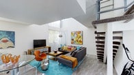 This dynamic two-story space makes a striking first impression with its soaring double-height ceilings in the main living area, creating an airy, expansive atmosphere that is both dramatic and inviting. Large windows allow generous natural light to flood the interior, enhancing the bright white walls and light-toned tile flooring that run throughout the main level.

The living area is thoughtfully designed with a mid-century modern aesthetic, anchored by a plush sectional sofa in charcoal gray accented with vibrant throw pillows in teal, mustard, and pink. A bold, colorful abstract artwork serves as a focal point above the sofa, complemented by an arc floor lamp and a sleek wooden media console. A vivid turquoise area rug adds warmth and energy underfoot, while an iconic orange accent chair contributes a playful pop of color.

A staircase with a distinctive spiral-wrapped column connects the main level to the upper floor, serving as both a functional and architectural feature. The upper level overlooks the living area below, contributing to the sense of vertical openness that defines this space.

Overall, the space combines bold, expressive design choices with a well-organized layout, offering a lively and contemporary environment that is visually engaging and rich in character.