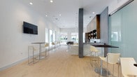 This single-floor space showcases a refined contemporary design centered around an open-plan layout that feels expansive and airy. Light hardwood flooring runs throughout, complementing the bright white ceiling fitted with recessed lighting that ensures consistent illumination across the entire area. A series of large windows and glass doors at the far end flood the interior with natural light and provide views of an outdoor area beyond.

The space is anchored by a striking wet bar featuring a marble-look countertop and warm wood cabinetry with open shelving for display. A dark countertop bar with modern cream-toned high chairs sits adjacent, creating an intimate seating area. Additional bar-height tables and matching chairs are arranged throughout, offering flexible gathering spots.

Architectural interest is provided by a prominent concrete column that adds an industrial contrast to the otherwise polished interior. A wall-mounted flat-screen television is positioned for easy viewing from multiple seating areas. Pendant lighting elements hang from the ceiling, adding subtle design accents. The overall ambiance is clean, minimalist, and functional, making it well-suited as a lounge or social amenity space.