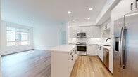 This contemporary single-floor residence showcases a clean, modern aesthetic with a cohesive palette of crisp whites, light wood flooring, and matte black hardware accents carried consistently throughout the space. The open-concept kitchen serves as a central highlight, featuring flat-front white cabinetry, a striking marble-veined quartz countertop island, a matching backsplash, stainless steel appliances including a gas range, microwave, dishwasher, and French door refrigerator, all illuminated by recessed ceiling lighting. The kitchen flows seamlessly into the living area, creating an expansive, airy feel ideal for both everyday living and entertaining. The primary bathroom offers a sophisticated double-vanity setup with a wide-format framed mirror, quartz countertops, and matte black fixtures. A secondary full bathroom provides a single vanity, a tub-shower combination, and tile flooring. The bedrooms are bright and well-proportioned, with large windows allowing natural light and views of the surrounding neighborhood. One bedroom features built-in shelving and a small built-in cabinet, adding functional storage. A dedicated walk-in closet with built-in rod systems serves the primary bedroom. Completing the unit is a convenient in-unit stacked washer and dryer tucked behind a louvered door.