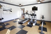 This fitness center offers a comprehensive workout environment designed to support a wide range of exercise routines. The space features a two-toned rubber tile floor in a classic checkerboard pattern of tan and black, providing both durability and cushioning underfoot. Ceiling fans with warm-toned light fixtures are mounted throughout, ensuring consistent airflow and comfortable ambient lighting alongside recessed ceiling lights.

The gym is outfitted with a solid lineup of equipment, including a treadmill, an elliptical cross-trainer, a multi-station cable weight machine, and a dumbbell rack stocked with a full range of free weights. A wall-mounted flat-screen television is positioned for easy viewing during cardio sessions. A large wall mirror spans one side of the room, visually expanding the space and allowing users to monitor their form during workouts.

Natural light streams in through multiple windows dressed with horizontal blinds, and the views beyond reveal an outdoor pool area. Multiple glass-paneled exit doors provide direct access to the exterior. The walls are painted in a clean light gray with bold dark gray trim, contributing to a modern, polished aesthetic. A piece of abstract wall art adds a subtle decorative touch, balancing the functional atmosphere of the fitness center.