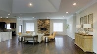 This single-floor space offers a thoughtfully designed interior that blends comfort with refined style. The main living area serves as the centerpiece, showcasing rich, dark hardwood flooring that extends throughout and adds warmth to the overall aesthetic. A striking stacked stone fireplace rises as a dramatic focal point, providing both visual interest and a cozy atmosphere. The tray ceiling above lends an elevated sense of height and architectural character to the room.

Recessed lighting is strategically placed throughout the ceiling, complementing the generous natural light that floods in through multiple white-trimmed windows dressed with clean, classic blinds. The neutral white and gray wall tones create a bright, airy backdrop that allows furnishings and architectural details to stand out.

The layout is open and spacious, accommodating a comfortable seating arrangement that includes soft upholstered sofas and accent chairs in warm yellow tones, paired with a rustic wooden coffee table. Crown molding along the upper walls adds a polished finishing touch. The overall ambiance is clean, modern, and inviting — a well-appointed space that balances architectural detail with functional, elegant design.