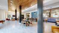 This contemporary single-floor clubroom is designed as a multifunctional community lounge, seamlessly blending social, dining, and relaxation zones within one open-plan layout. The space immediately draws the eye upward with its striking warm-toned wood slat ceiling, which contrasts beautifully against crisp white walls and sleek dark steel columns that punctuate the floor plan. Underfoot, large-format marble-look tile flooring runs throughout, lending a clean, polished aesthetic to every area.

The dining zone features round black tables paired with mid-century modern wooden chairs, creating an inviting setting for casual gatherings. A built-in banquette upholstered in soft periwinkle blue is flanked by bistro-style tables and illuminated by gold dome pendant lights, with a large mirror panel above amplifying the sense of space. Adjacent to the banquette, a textured blue scallop-patterned tile accent wall anchors a wet bar or café counter with warm wood cabinetry.

The lounge area is anchored by a statement circular chandelier with warm-toned bulbs set within a wood-trimmed tray, positioned above a curated seating arrangement of a caramel leather sofa, rust-orange accent chairs, and a tufted leather ottoman centered on a layered area rug. A wall-mounted flat-screen TV completes this entertainment-ready retreat. Floor-to-ceiling windows throughout flood the space with natural light and offer views of the surrounding outdoor pool and courtyard.