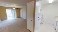 This single-floor residence offers a straightforward, functional layout designed for comfortable everyday living. The open-concept design seamlessly connects the living area and kitchen, creating a cohesive and practical flow throughout the space. The living area is finished with wall-to-wall carpeting and features a ceiling fan, providing year-round comfort. Large vertical-blind-covered sliding glass doors allow natural light to fill the room while offering access to the outdoors. The kitchen is situated adjacent to the living space and is equipped with white cabinetry, white appliances including a refrigerator, a cooktop range, and ample counter space. The kitchen's light-toned flooring contrasts neatly with the carpeted living area, clearly delineating the two zones. A full-height pantry or storage closet is positioned at the transition between the kitchen and living area, providing convenient organizational space. The interior walls are finished in neutral warm tones throughout, contributing to a clean, understated ambiance. The front entry door is visible from the main living area, reinforcing the open sightlines of the layout. Overall, the space presents a practical and well-organized residential environment suited for a range of occupants.