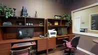 This single-floor business center is designed to provide a practical and comfortable workspace for those needing to stay connected and productive. The room features two side-by-side wooden workstation units, each equipped with a desktop computer and monitor, offering ample desk surface for working, browsing, or printing. A multifunction printer/scanner sits conveniently between the two stations, ensuring easy access from either workstation. Above each desk, open shelving units provide additional storage and display space, decorated with potted greenery and coffee-themed accents that lend the room a welcoming, informal ambiance. The warm-toned wood cabinetry contrasts with the deep brown walls, creating a cozy yet focused atmosphere. Ergonomic task chairs accompany each workstation, supporting extended periods of use. Natural light filters in through a nearby window, brightening the space without overwhelming it. The hardwood flooring adds a polished, professional feel to the overall aesthetic. Whether used for business correspondence, travel planning, or general browsing, this well-equipped business center offers everything needed for a convenient and efficient work session.