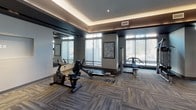 This contemporary fitness center is thoughtfully designed across a single floor, offering a comprehensive workout environment suited for a wide range of exercise routines. The space is organized into distinct functional zones, each catering to different training needs. Large windows throughout the facility flood the interior with natural light and provide views of the surrounding exterior, creating an open and energizing atmosphere.

The strength training area features a full dumbbell rack, adjustable weight bench, and a barbell squat rack, all set against bold green accent walls and floor-to-ceiling mirrors that enhance the sense of space. Pendant lighting adds a stylish touch overhead. A dedicated cardio zone houses stair climbers, stationary bikes, and rowing machines, positioned near expansive windows to maximize natural light during workouts.

A functional training area provides open floor space equipped with plyo boxes, kettlebells, and a utility bench, ideal for dynamic and high-intensity training. Mirrored walls throughout multiple zones support form-checking during exercise. The flooring is consistent throughout, featuring durable, patterned carpet tiles in neutral tones with subtle color accents. A tray ceiling with indirect lighting in one section adds architectural depth. The overall design is modern and cohesive, balancing aesthetics with practical fitness functionality.