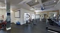 This single-floor fitness center is thoughtfully designed to support a full range of workout routines. The space features a durable rubber flooring in a dark speckled finish, providing both safety and functionality underfoot. Overhead, an elegant coffered ceiling with recessed lighting adds architectural character, while a ceiling fan ensures comfortable air circulation during workouts.

The gym is outfitted with a variety of strength training equipment, including cable machines, a multi-functional weight station, and a well-stocked dumbbell rack, catering to users of varying fitness levels. A large wall-mounted projection screen on one end of the room offers entertainment or instructional viewing during exercise sessions. Natural light filters in through multiple windows fitted with blinds, brightening the space and creating an inviting atmosphere.

A floor-to-ceiling mirror near the entrance enhances the sense of openness and allows users to monitor their form. The accent stonework around the entryway adds a refined, upscale aesthetic to the space. A stability ball and additional accessories round out the available equipment. Overall, the fitness center combines practical amenities with polished design elements to deliver a comfortable and motivating environment for exercise.