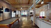 This expansive single-floor space is a masterfully converted industrial building in South Carolina, originally part of the Woodside Fine Fabrics facility—a vintage circular logo still proudly displayed on the original brick wall serves as a nod to its storied past. The open-plan layout is defined by soaring ceilings clad in reclaimed wood planks supported by heavy timber beams and original wooden columns, while exposed ductwork and polished concrete floors reinforce the space's authentic industrial character.

The layout flows seamlessly between distinct functional zones. A spacious game area anchors one end, featuring a full-size pool table beneath statement pendant lighting and a wall-mounted television, alongside original corrugated metal sliding doors and large glazed entry doors that flood the space with natural light. The living area offers generous seating, hanging egg chairs, an oversized area rug, a large-blade ceiling fan, and another wall-mounted television set against raw brick. At the far end, a modern kitchen with a backlit tile backsplash, a large island with bar seating, pendant lighting, and stainless steel appliances provides a refined contrast to the rustic surroundings. Dining areas with wooden tables complete the space.