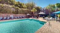 Located at 1100 Pulaski St in Columbia, South Carolina, this outdoor amenity space offers a well-appointed retreat designed for relaxation and social gathering. The centerpiece is a generously sized, crystal-clear swimming pool set within a brick-trimmed surround, flanked by rows of lounge chairs and shaded umbrella seating that invite extended outdoor leisure. The pool area is enclosed by mature trees and manicured hedgerows, providing a sense of privacy while maintaining an open, airy atmosphere under the abundant South Carolina sunshine. An adjacent brick clubhouse structure with arched windows and French doors serves as a functional anchor to the pool deck, offering convenient access to indoor amenities. Beyond the pool, a separate patio area features a stone fire pit encircled by contemporary grey sectional seating with teal cushions, creating an inviting gathering space ideal for evening socializing. The surrounding landscape is thoughtfully designed with colorful shrubs, ornamental grasses, decorative river rock, and swaying palm trees that lend a resort-like tropical ambiance. The combination of brick architecture, paved stone pathways, and lush greenery gives the overall space a cohesive, upscale aesthetic well-suited to the vibrant Columbia community.