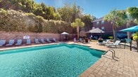 Located at 1100 Pulaski St in Columbia, South Carolina, this outdoor amenity space offers a well-appointed retreat designed for relaxation and social gathering. The centerpiece is a generously sized, crystal-clear swimming pool set within a brick-trimmed surround, flanked by rows of lounge chairs and shaded umbrella seating that invite extended outdoor leisure. The pool area is enclosed by mature trees and manicured hedgerows, providing a sense of privacy while maintaining an open, airy atmosphere under the abundant South Carolina sunshine. An adjacent brick clubhouse structure with arched windows and French doors serves as a functional anchor to the pool deck, offering convenient access to indoor amenities. Beyond the pool, a separate patio area features a stone fire pit encircled by contemporary grey sectional seating with teal cushions, creating an inviting gathering space ideal for evening socializing. The surrounding landscape is thoughtfully designed with colorful shrubs, ornamental grasses, decorative river rock, and swaying palm trees that lend a resort-like tropical ambiance. The combination of brick architecture, paved stone pathways, and lush greenery gives the overall space a cohesive, upscale aesthetic well-suited to the vibrant Columbia community.