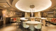 This single-floor space exudes refined elegance through its thoughtfully curated design and upscale finishes. At the heart of the space is a grand, curved bar finished in a soft neutral palette, surrounded by plush upholstered bar stools that invite guests to settle in comfortably. The bar's sweeping horseshoe shape creates an intimate yet expansive focal point that anchors the entire room.

Above, a dramatic coffered ceiling features a circular recessed tray with soft ambient lighting, complemented by a stunning cluster of glass pendant lights that cascade at varying heights, adding a jewel-like quality to the space. Recessed lighting throughout ensures the room is evenly illuminated while maintaining a warm, inviting atmosphere.

Along the walls, an intricate large-scale botanical mural serves as a striking artistic backdrop, adding depth and a touch of nature-inspired elegance to the refined interior. A cozy lounge seating area with accent pillows in rich orange tones is visible in the background, offering a relaxed alternative to bar seating.

The overall design language blends classic and contemporary elements — from the neutral upholstery and warm wood tones to the decorative wall panels — creating a cohesive, luxurious environment well-suited for sophisticated social gatherings.