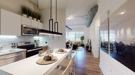 Located at 1 Martine Ave in White Plains, NY, this modern loft-style residence showcases an open-concept layout defined by dramatic soaring ceilings with exposed ductwork and industrial-style track lighting, all grounded by rich hardwood flooring throughout. The kitchen is a functional and stylish centerpiece, featuring two-tone cabinetry with warm wood lower cabinets and sleek white upper cabinets, a subway tile backsplash, stainless steel appliances, under-cabinet lighting, and a white waterfall island with bar seating. A bold geometric pendant light anchors the dining area just beyond, which flows seamlessly into the living room. The living room is bright and airy, with large windows offering views of the surrounding White Plains cityscape, track lighting overhead, and generous wall space for artwork. The bedroom is a serene retreat with hardwood floors, soft neutral tones, exposed ceiling beams, and a window with natural light. A dedicated home office nook provides built-in wood cabinetry and desk space. The bathroom is finished with large-format tile flooring, a wood-front vanity, and a well-lit mirror. Throughout, the space balances industrial architecture with warm, contemporary design elements for a cohesive and inviting ambiance.