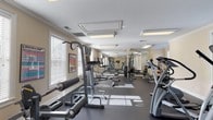 Located at 100 Legends Dr in Montgomery, Alabama, this fitness center offers a well-appointed, single-floor workout facility designed to support a full range of exercise needs. The space features warm beige walls, elegant crown molding, and a dark rubber flooring that provides both durability and comfort underfoot. Recessed fluorescent lighting panels illuminate the room evenly, while multiple large windows fitted with white horizontal blinds allow ample natural light to filter in, creating a bright and energizing atmosphere. A full-length mirrored wall along one side visually expands the space and aids in proper form during workouts. The cardio section is outfitted with multiple treadmills, elliptical machines, and a stationary bike, while the strength training area includes a cable machine, weight benches, barbells, and a well-stocked dumbbell rack. A mounted flat-screen television is positioned for convenient viewing during workouts, and a fitness reference chart is displayed on the wall for guidance. The facility is accessed through a glass-paned door and includes safety features such as a visible fire extinguisher and emergency exit signage. The overall ambiance is clean, functional, and welcoming, making it a practical and accessible fitness resource.