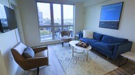 This single-floor living space offers a contemporary and inviting atmosphere, centered around a generously sized living area. The standout feature is the expansive floor-to-ceiling windows that flood the room with natural light while framing sweeping views of the surrounding cityscape and what appears to be a marina in the distance. A private balcony, accessible through the large window wall, extends the living area outdoors and provides an ideal spot for enjoying the views.

The living room is finished with light-toned hardwood flooring that adds warmth to the space. Furnishings are thoughtfully arranged around a geometric patterned area rug, anchoring a white oval coffee table with gold-toned legs. Seating includes a rich blue sofa adorned with accent pillows, a tan leather armchair, and a floral accent chair positioned near the windows to take advantage of the natural light and views. A framed blue artwork on the wall complements the color palette of the room.

The overall ambiance is modern and comfortable, with a neutral wall palette that allows the furnishings and natural light to serve as the primary design elements, creating a welcoming and well-curated living environment.