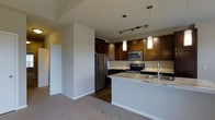 This two-story residence offers a thoughtfully designed layout that balances functionality with modern style. The kitchen serves as a standout feature, showcasing rich dark wood cabinetry that extends to the upper walls, providing generous storage throughout. A peninsula with elegant granite countertops — featuring warm tones and natural veining — anchors the space and offers additional prep and seating areas. Stainless steel appliances, including a refrigerator, electric range, and over-the-range microwave, lend a sleek, contemporary feel. Pendant lighting with cylindrical white shades hangs above the peninsula, complemented by a track lighting system that illuminates the full kitchen area. The flooring transitions from hardwood in the kitchen to plush carpet in the adjacent living area, creating a subtle distinction between spaces. Neutral wall tones throughout contribute to a clean, open ambiance. The two-floor layout provides versatile living across multiple levels, making the space well-suited for a variety of lifestyles. Overall, the interior strikes a balance between practical design and refined finishes.