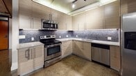 This single-floor urban loft showcases a compelling blend of industrial character and contemporary finishes throughout its thoughtfully designed layout. Polished concrete floors run through the open living areas, complementing the raw, industrial aesthetic defined by exposed ceiling pipes, track lighting, and steel-framed factory-style windows that allow natural light to pour in and offer views of the surrounding urban environment.

The kitchen is a standout feature, outfitted with a full suite of stainless steel appliances including a range with over-the-range microwave, side-by-side refrigerator, and dishwasher. Light wood-grain flat-front cabinetry lines the walls from floor to ceiling, accented by a striking blue mosaic tile backsplash and under-cabinet LED lighting. The countertops are clean and bright, providing ample workspace.

The bathroom presents a refined, modern aesthetic with large-format gray floor tiles, a floating wood-tone vanity topped with a vessel sink, and a combined soaking tub and shower enclosed by sleek frameless glass doors. Deep charcoal stacked tile surrounds the tub, adding depth and contrast against the crisp white walls.

The living area is open and versatile, featuring polished concrete flooring, industrial-style exposed ceiling elements, and a frosted glass sliding barn door that leads to a closet or secondary space. The expansive windows with curtain panels complete the loft's urban-industrial ambiance.
