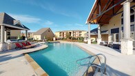This single-story amenity space is designed to support an active lifestyle, combining a fully outfitted fitness center with an inviting outdoor pool area. The fitness center is organized across several interconnected zones, each serving a distinct workout purpose. Cardio equipment is abundantly represented, with a long row of treadmills, ellipticals, stationary bikes, and recumbent bikes lining the carpeted floor alongside large windows that allow generous natural light to fill the space. Mirrors along the walls visually expand the room and aid in form monitoring during exercise. A dedicated strength-training section features a Life Fitness cable machine, a power rack with a flat bench, free weights, and various attachments, while an adjacent area stores stability balls and supplementary fitness accessories. A wall-mounted television, a water cooler, and towel storage contribute to user convenience. A glass-partitioned room with vaulted ceilings and arched windows provides a separate studio space for stretching or group activities. Outdoors, the resort-style pool features a broad, curved design with clear turquoise water, chrome entry rails, and a generous surrounding deck furnished with lounge chairs. A covered pavilion with wood-accented ceilings and open-air columns provides shaded seating adjacent to the pool.