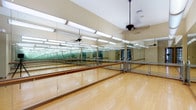 This yoga and Pilates studio is purpose-built for movement-based fitness, offering a spacious, open floor plan with a clean and functional aesthetic. The studio's most defining feature is its expansive floor-to-ceiling mirrored walls, which line multiple sides of the room, creating a sense of depth and allowing practitioners to monitor their form with precision. Suspended fluorescent lighting fixtures run in parallel rows across the ceiling, providing bright, even illumination throughout the space.

The flooring consists of smooth, polished hardwood in a warm honey tone, offering a resilient yet forgiving surface ideal for barefoot practice. Multiple rows of freestanding and wall-mounted ballet barres are positioned throughout the studio, supporting barre-based yoga, Pilates, and stretching exercises. Ceiling fans are installed overhead, ensuring consistent airflow and comfort during active sessions.

Natural light enters through glass-paneled double doors along one wall, connecting the studio to an adjacent corridor and adding a welcoming openness to the environment. The neutral warm beige walls complement the hardwood floors, contributing to a calm and focused atmosphere well-suited for both individual practice and group instruction.