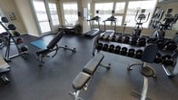 This fitness center is a thoughtfully designed single-floor space that combines functionality with an inviting atmosphere. Large windows line the walls on multiple sides, flooding the interior with natural light while providing panoramic views of a nearby waterway and surrounding landscape — creating an energizing backdrop for workouts.

The flooring throughout consists of durable dark rubber matting, a practical choice that provides cushioning and stability for a range of exercise activities. The strength training area is well-appointed, featuring multiple adjustable weight benches and an extensive dumbbell rack stocked with a full range of free weights, accommodating users of varying fitness levels.

The cardio section includes multiple treadmills positioned directly in front of the large windows, allowing users to enjoy the waterfront scenery during their workouts. Additional strength and resistance equipment is visible along the perimeter of the room, making efficient use of the available space.

The neutral-toned walls complement the overall clean, professional aesthetic of the facility. A decorative plant and framed artwork add subtle finishing touches, balancing the utilitarian nature of the equipment with a welcoming ambiance.