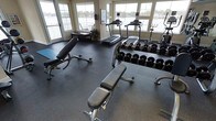 This fitness center is a thoughtfully designed single-floor space that combines functionality with an inviting atmosphere. Large windows line the walls on multiple sides, flooding the interior with natural light while providing panoramic views of a nearby waterway and surrounding landscape — creating an energizing backdrop for workouts.

The flooring throughout consists of durable dark rubber matting, a practical choice that provides cushioning and stability for a range of exercise activities. The strength training area is well-appointed, featuring multiple adjustable weight benches and an extensive dumbbell rack stocked with a full range of free weights, accommodating users of varying fitness levels.

The cardio section includes multiple treadmills positioned directly in front of the large windows, allowing users to enjoy the waterfront scenery during their workouts. Additional strength and resistance equipment is visible along the perimeter of the room, making efficient use of the available space.

The neutral-toned walls complement the overall clean, professional aesthetic of the facility. A decorative plant and framed artwork add subtle finishing touches, balancing the utilitarian nature of the equipment with a welcoming ambiance.