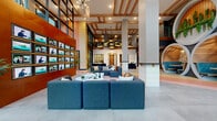 Located at 1015 N Alexander Street in Charlotte, NC, this single-floor communal space is a masterclass in contemporary interior design. The entrance opens into a generously proportioned lobby and lounge area defined by soaring double-height ceilings with exposed wooden beam detailing, creating an open and airy atmosphere. Structural columns clad in bold checkerboard and white tile patterns serve as eye-catching architectural accents throughout the space.

The flooring is one of the most distinctive features, showcasing a dynamic geometric pattern in shades of yellow, coral, pink, and grey that delineates different zones across the open floor plan. Modular grey sofas and lounge seating are thoughtfully arranged to encourage gathering and conversation.

A standout feature is a cluster of large circular tunnel pods — finished with warm wooden slat interiors, colorful mosaic tile backsplashes, and teal booth seating — that offer intimate, semi-private nooks within the larger open space. Live greenery cascades from the tops of these pods, adding a natural, organic element.

A wall of mounted screens on warm wood paneling anchors one side of the room, while floor-to-ceiling windows and glass doors connect the interior to an outdoor area with views of a covered patio. A bar-height counter with orange stools sits adjacent to the outdoor access, enhancing the space's social and functional appeal.