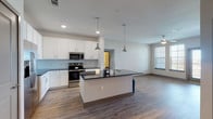 This single-floor residence offers three bedrooms and two bathrooms within a thoughtfully designed open-concept layout. The kitchen serves as a central hub, featuring white shaker cabinetry, a light gray subway tile backsplash, dark granite countertops, and a full suite of stainless steel appliances including a range, microwave, and refrigerator. A large center island with a dark countertop and integrated sink provides additional prep space and anchors the room, illuminated by a pair of industrial-style pendant lights. Wood-look flooring extends throughout the main living areas, creating a cohesive and easy-to-maintain aesthetic.

The open living area adjoins the kitchen seamlessly, benefiting from natural light through multiple windows and a ceiling fan overhead. The bedrooms are carpeted for comfort, with at least one room featuring impressively tall windows that reach toward the ceiling, offering expansive outdoor views and abundant daylight. Additional bedrooms provide well-proportioned, versatile spaces.

Both bathrooms are finished with white vanities, dark granite countertops, and wood-look tile flooring. One bathroom includes a full tub-shower combination with white subway tile surround, while the second bathroom features a separate shower area. The overall design maintains a clean, contemporary palette throughout.