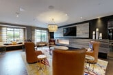 Located at 70 Upper Rock Cir in Rockville, Maryland, this thoughtfully designed communal lounge space offers a refined and inviting atmosphere across a single floor. The living room serves as the focal point of the space, anchored by a striking floor-to-ceiling stone-clad wall housing a wide linear gas fireplace, above which a large flat-screen television is mounted. Rich hardwood flooring runs throughout, complementing the warm, earthy tones of the interior palette. Plush cognac and burgundy leather armchairs are arranged in conversation groupings atop vibrant, patterned area rugs, creating intimate seating clusters suited for both socializing and relaxation. A tiered drum chandelier with warm ambient lighting adds an elegant touch overhead, supplemented by recessed ceiling lights throughout. Natural light filters in through multiple windows dressed with striped roller shades, offering views of the surrounding greenery. A communal work or dining table with bar-height seating is visible toward the rear, illuminated by a series of colorful pendant lights. Glass partition walls define a more private seating nook, adding an architectural layer of openness and transparency. The overall ambiance blends modern sophistication with warm, residential comfort.