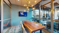 This well-appointed amenity lounge is a thoughtfully designed communal space that seamlessly blends multiple functional zones within a single, open floor plan. The interior is unified by a warm hardwood floor and a consistent teal color palette across textured walls, creating a cohesive and inviting atmosphere throughout.

A central highlight is the long communal dining table crafted from rich, dark wood, surrounded by upholstered bar-height chairs and anchored above by a striking geometric lantern-style chandelier. Adjacent to this gathering area, a smaller round table with elegantly detailed teal chairs sits beside a glass door that opens directly to an outdoor terrace, filling the corner with natural light and greenery views.

A dedicated lounge area features a curated arrangement of plush sofas, swivel accent chairs in soft aqua tones, and a low-profile coffee table, all oriented around a wall-mounted television and complemented by framed artwork. Floor-to-ceiling windows along one wall flood this seating zone with abundant natural light while offering panoramic views of an outdoor courtyard and the surrounding cityscape.

Decorative lattice screens serve as elegant architectural dividers between zones, adding visual depth without sacrificing openness. High bistro tables with gold-accented chairs provide additional flexible seating. The overall ambiance is polished yet comfortable, balancing contemporary design elements with warm, welcoming furnishings ideal for both socializing and relaxing.