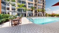 Located in Miami, FL, this outdoor pool deck is a vibrant amenity space designed to capture the essence of South Florida resort living. The centerpiece is a generously sized swimming pool with crystal-clear turquoise water, framed by clean tile edges and accessible via stainless steel handrails. Surrounding the pool, a broad paved deck provides ample space for relaxation, lined with rows of cushioned chaise lounges shaded by bold red market umbrellas and pergola structures. An adjacent outdoor dining area features sturdy table-and-chair sets, ideal for al fresco gatherings. Lush tropical landscaping, including tall swaying palm trees, manicured hedgerows, flowering shrubs, and ground-level pathway lighting, creates a verdant border that adds privacy and a resort-like ambiance. The deck is set within an interior courtyard formed by a multi-story residential building, whose contemporary façade — accented in contrasting shades of orange and gray — rises dramatically on all sides. The open-sky setting allows abundant natural sunlight to flood the space throughout the day, while the surrounding balconies offer elevated views down to the pool area below.