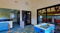 This single-floor amenity space is thoughtfully designed to support both recreation and fitness. The layout opens into a bright, airy game room distinguished by soaring ceilings and large black-framed windows that allow natural light to filter throughout. A colorful striped area rug anchors the space and adds a lively, energetic feel to the room. A foosball table serves as a central recreational feature, making the area ideal for social interaction and casual play.

Black-framed glass doors connect the game room to the adjacent fitness center, which is visible through large windows and provides a seamless visual connection between the two areas. The fitness center is well-equipped with a variety of exercise machines, including weight and cardio equipment, set against vibrant walls that create an energizing workout environment.

The neutral, light-toned walls throughout the space complement the bold accents of color found in the furnishings and flooring, striking a balance between functionality and visual interest. Ventilation is well-integrated into the ceiling, contributing to a comfortable atmosphere across all areas. Overall, this amenity space offers a well-rounded environment that caters to both active recreation and structured fitness activity.