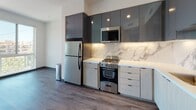 This contemporary single-floor unit showcases a thoughtfully designed open layout with consistent dark hardwood-style flooring throughout. The kitchen is a standout feature, outfitted with high-gloss charcoal upper cabinetry, light wood lower cabinets, and pristine white quartz countertops. A striking marble-look backsplash runs the full length of the cooking area, complementing the suite of stainless steel appliances that includes a gas range, built-in microwave, full-size refrigerator, and dishwasher. Recessed under-cabinet lighting adds both function and ambiance to the culinary space. The living area benefits from generous natural light, with floor-to-ceiling glass panels and a sliding door that opens directly onto a private balcony, offering open views of the surrounding neighborhood. The bathroom is finished in a clean, minimalist style featuring warm wood-toned tile surround on the tub-shower combination, a wide white vanity with undermount sink, and wood-grain vinyl flooring that ties the space together cohesively. Throughout the unit, the bright white walls and modern finishes create a fresh, airy atmosphere well-suited to contemporary urban living.