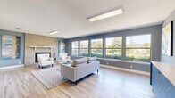 This resident lounge in Austin, TX offers a well-appointed communal space designed for comfort and social gathering. The living area is the centerpiece of the lounge, featuring light hardwood-style flooring, soft blue-gray walls, and an eye-catching brick fireplace with a white mantel that serves as a focal point. A row of large windows lines one wall, flooding the room with abundant natural light and providing pleasant views of the surrounding greenery and outdoor landscape. The seating area is thoughtfully arranged with a contemporary tufted sofa, cream-colored armchairs, a glass-and-gold coffee table, and a patterned area rug, creating an inviting and cohesive aesthetic. Colorful abstract artwork adorns the walls, adding a vibrant, modern touch to the space. Recessed lighting and flush-mount ceiling fixtures ensure the room remains well-lit throughout the day. Adjacent to the living area, a dedicated kitchen features rich dark wood cabinetry, granite countertops, a full-size refrigerator, a microwave, and a Keurig coffee maker — providing residents with convenient amenities for everyday use. The tile flooring in the kitchen contrasts neatly with the living area, clearly delineating the two functional zones within this comfortable and inviting lounge.