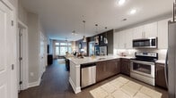 This well-appointed single-floor residence offers a thoughtfully designed open-concept layout that seamlessly connects the kitchen, dining, and living areas. The kitchen is a standout feature, equipped with stainless steel appliances including a range, microwave, and dishwasher, complemented by a two-tone cabinet scheme pairing dark espresso lower cabinets with crisp white upper cabinets. Granite countertops extend across the surfaces and onto the kitchen island, which is illuminated by a series of stylish pendant lights. A subway tile backsplash adds a refined touch throughout. Dark hardwood flooring runs through the main living areas, anchoring the open-concept space. The living room features an accent wall with textured wallcovering, large windows that invite generous natural light, and a ceiling fan overhead. Two bedrooms are present, each with large windows offering outdoor views. One bedroom features a textured accent wall, while the other showcases bold, dark-painted walls for contrast. A spacious walk-in closet with wire shelving and built-in hanging rods provides ample storage. The bathroom is finished with large-format tile flooring, a granite vanity with dark cabinetry, and a glass-enclosed shower, offering a clean and functional design throughout.