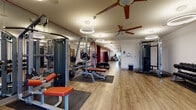 Located on Hayes Court in Beavercreek, Ohio, this fitness center offers a comprehensive, single-floor exercise facility designed to accommodate a wide range of workout needs. The space is organized into distinct zones, separating strength training and cardio areas while maintaining an open, unobstructed flow throughout.

The strength training section is well-appointed with multiple cable machines, weight benches, barbells, plate-loaded equipment, and a dumbbell rack. Full-length mirrors line portions of the walls, supporting proper form during workouts. Rubber floor mats are positioned beneath equipment to protect the wood-look luxury vinyl flooring that extends throughout the facility.

The cardio zone features Matrix-brand machines, including treadmills, ellipticals, a stair climber, and rowing machines, providing users with a variety of aerobic training options. A sanitizing station is conveniently located within this area.

Overhead, circular ring pendant lights provide bright, even illumination across the entire space, complemented by ceiling fans that promote air circulation. Bold terracotta-red accent walls create visual contrast against the neutral white walls and light flooring. A window at the far end of the space allows natural light to filter in, enhancing the overall ambiance. The facility also includes a separate door providing access to an adjacent room.