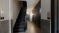 Located at 2054 Avenue Claremont in Montréal, this building entrance presents a well-appointed and thoughtfully designed common area comprising two interconnected hallways. The space makes a strong first impression through its blend of classic architectural elements and refined modern finishes.

The primary hallway is anchored by a striking staircase featuring dark-painted wooden steps, a rich warm-toned handrail, and a prominent newel post, conveying a sense of heritage character. One wall is clad in deep charcoal-toned wainscoting with framed panel molding, upon which a framed artwork is displayed, lending the corridor a polished, gallery-like quality. The floor throughout is laid in large-format neutral stone-effect tiles, offering a clean and durable surface.

The secondary hallway leads to the main building entrance door, which features a glazed window panel that allows natural light to filter in. An elevator is conveniently positioned along this corridor, ensuring accessibility throughout the building. Dark-framed interior doors provide visual continuity and contrast against the light grey upper walls and charcoal lower tile cladding.

Wall-mounted sconce lighting contributes to a warm, welcoming ambiance. Safety equipment, including a fire extinguisher, is discreetly integrated. Together, the two hallways form a functional yet characterful entrance that effectively balances the building's historic architectural roots with contemporary upkeep.