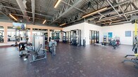 This well-appointed fitness center in Phoenix, AZ offers a spacious, single-floor layout divided into two distinct exercise rooms designed to accommodate a wide range of workout styles. The main exercise area features an industrial-inspired aesthetic with exposed steel trusses, open ductwork, and high ceilings that lend the space an open, airy feel. Large windows and glass garage-style doors flood the floor with abundant natural light while offering views of the surrounding exterior. The main floor is equipped with a comprehensive selection of strength training machines, cable systems, free weights including dumbbells and kettlebells, medicine balls, and a variety of cardio equipment such as stationary bikes, rowing machines, air bikes, and stair climbers. Durable rubber flooring throughout supports heavy use, while large wall mirrors enhance the sense of space. The second exercise room offers a dedicated studio environment with warm, recessed tray ceiling lighting, smooth finished walls, and hardwood-style flooring. This refined space is outfitted with pilates reformer machines and full-length mirrors, and is separated from the main floor by sleek glass partition doors, providing a quieter setting for focused training or group sessions. Oversized fans circulate air throughout both areas, ensuring comfort during intense workouts.