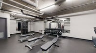 This fitness center is organized across a single level and divided into distinct training zones, offering a comprehensive workout environment for a variety of exercise styles. The strength training area is well-appointed with multiple adjustable weight benches, a full dumbbell rack spanning a wide range of weights, and a cable pulley machine, all positioned in front of a large wall-mounted mirror that enhances the sense of space and assists with form. A dedicated cardio zone features several treadmills and an elliptical trainer, backed by a vibrant wall mural of colorful athletic silhouettes that adds energy and visual character to the room. A functional training area provides open floor space alongside a medicine ball rack and a stability ball, offering room for dynamic and bodyweight exercises. The space also includes a rowing machine for additional cardio options. Throughout the facility, the ceiling is left exposed, showcasing concrete slabs, industrial piping, and ductwork, which lend the space a raw, urban aesthetic. Bright fluorescent lighting ensures consistent illumination across all zones. The durable black rubber flooring provides traction and impact absorption throughout. The overall atmosphere is utilitarian and purpose-driven, designed to support a full range of fitness activities in a compact, efficient layout.