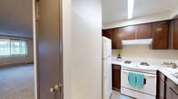 Located at 111 College Hill St in Cheney, WA, this single-floor unit offers a practical and well-organized layout across three bedrooms and a connecting hallway. The kitchen is efficiently designed with rich dark wood cabinetry, white appliances including a full-size refrigerator and electric range, a tile backsplash, and durable hard-surface flooring. Bright overhead lighting illuminates the workspace, making the kitchen both functional and inviting. The living area is carpeted and features a ceiling fan, baseboard heating, and neutral wall tones that create a clean, versatile backdrop for any furnishings. Each of the three bedrooms is carpeted and finished with neutral walls, dark wood trim accents, and textured ceilings. The generously sized primary bedroom includes a window with vertical blinds that brings in natural light and outdoor views, along with a wall-mounted air conditioning unit for added comfort. The two additional bedrooms are similarly appointed, each featuring windows with vertical blinds, baseboard heating, and ample floor space. A hallway connects the rooms, contributing to the efficient flow of the layout. Throughout the unit, consistent finishes and practical design elements provide a comfortable, functional living environment.
